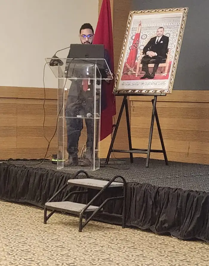 Le Dr. Achraf Ainouss, biologiste de l'Institut Pasteur de Casablanca, présente lors de la Journée de l'Amicale des ORL de Casablanca sur la tuberculose cervicale, avec en arrière-plan le drapeau marocain et un portrait du roi.